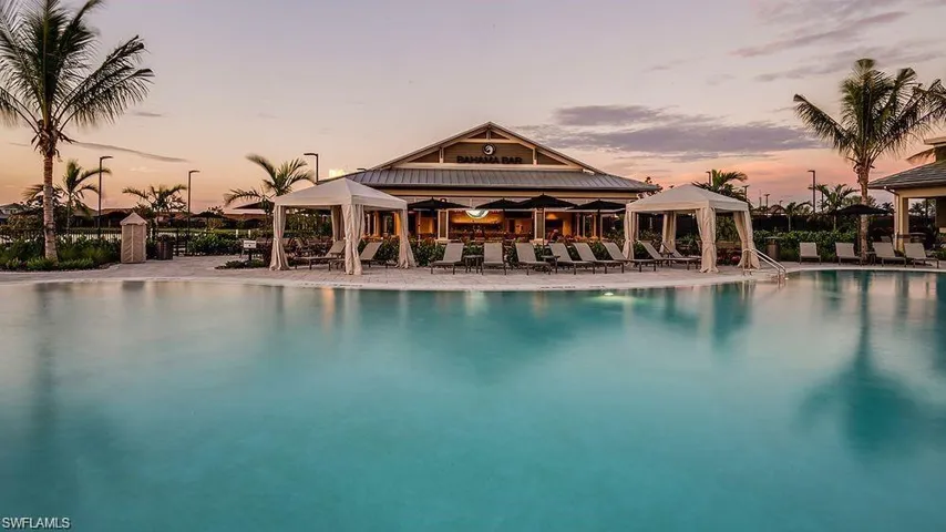 Resort-style swimming pool featuring a spacious deck, multiple cabanas, perimeter lounge seating, and a prominent clubhouse building with covered outdoor dining areas