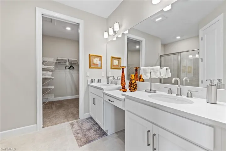 Spacious bathroom featuring a dual vanity with white cabinetry and light countertops, a large mirror with integrated lighting, and a walk-in closet with wire shelving