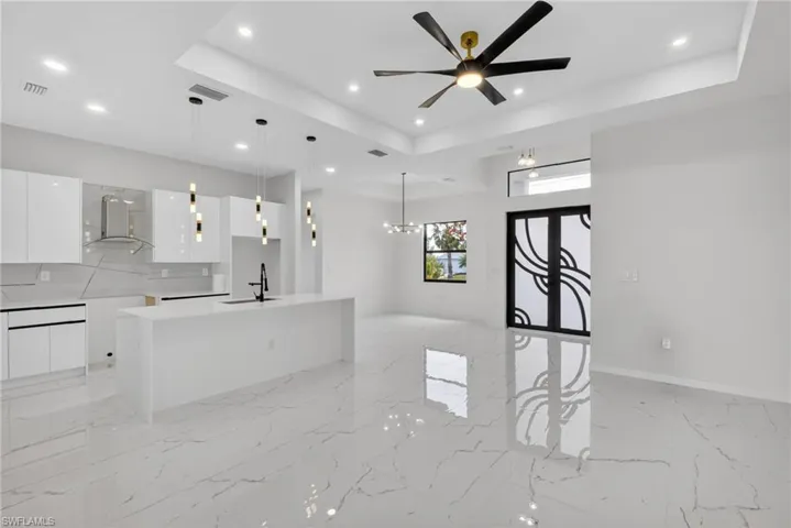 Open-concept living area featuring large format white tile flooring with veining, a contemporary kitchen island with pendant lighting, white high-gloss cabinetry, and a black ceiling fan with gold accents