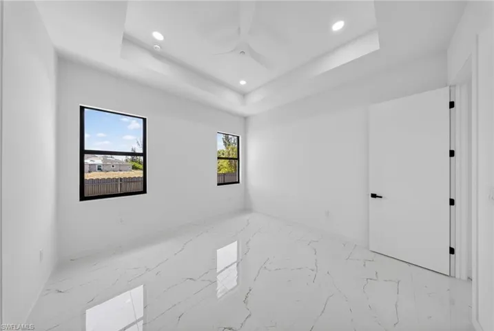 Spacious interior room featuring a tray ceiling with recessed lighting and a ceiling fan, white marble-look tile flooring, and two black-framed windows