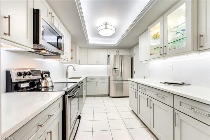 Kitchen with stainless steel appliances, a raised ceiling, glass fronted cabinets, light tile patterned floors, and light stone counters