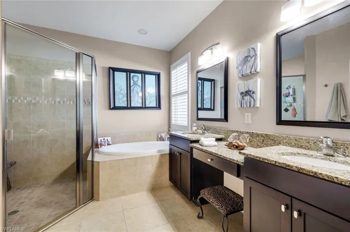 Spacious bathroom featuring a glass-enclosed shower, built-in soaking tub, dual vanities with granite countertops, and light-colored floor tiles