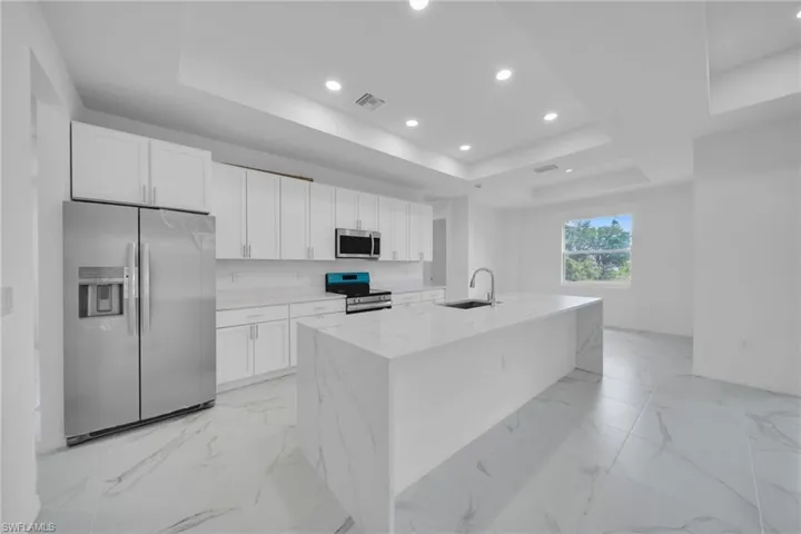 Spacious kitchen featuring an expansive island with waterfall countertops, stainless steel appliances, white shaker cabinetry, recessed lighting, and large format tile flooring