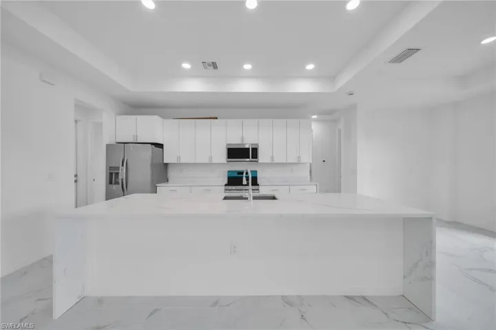 Modern kitchen featuring a large waterfall island with integrated sink, white cabinetry with silver hardware, stainless steel appliances, and recessed lighting