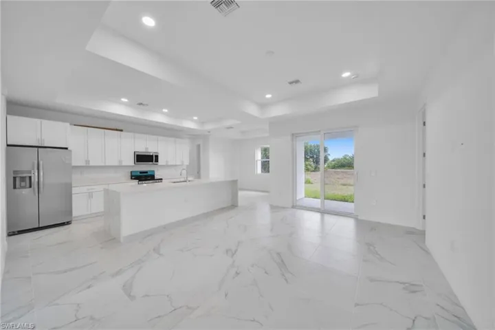 Open-concept living space featuring large format marble-look tile flooring, a kitchen with white cabinetry and stainless steel appliances, a center island with a sink, tray ceilings with recessed lighting, and sliding glass doors to the exterior