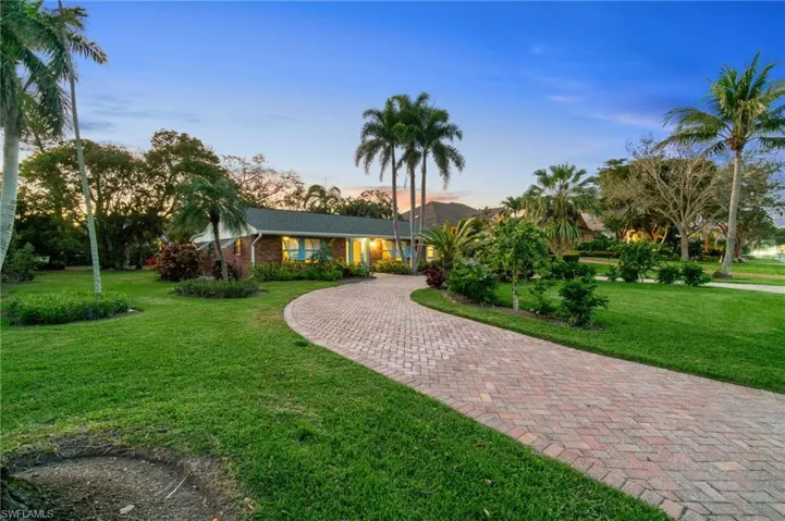 View of front of home with decorative paver circular driveway, brick siding, and a expansive yard with mature, tropical landscaping.