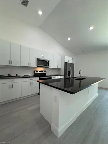 Kitchen with vaulted ceiling, a kitchen island with sink, white cabinets, stainless steel appliances, and light wood-style floors