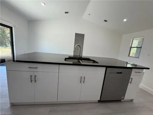Kitchen with stainless steel dishwasher, white cabinets, an island with sink, dark stone counters, and vaulted ceiling