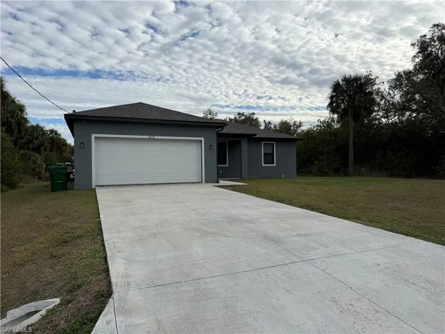 Ranch-style house featuring a front lawn, driveway, an attached garage, stucco siding, and roof with shingles