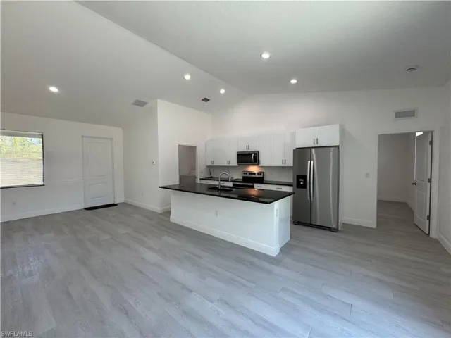 Kitchen featuring appliances with stainless steel finishes, a center island with sink, lofted ceiling, dark countertops, and light wood-style floors