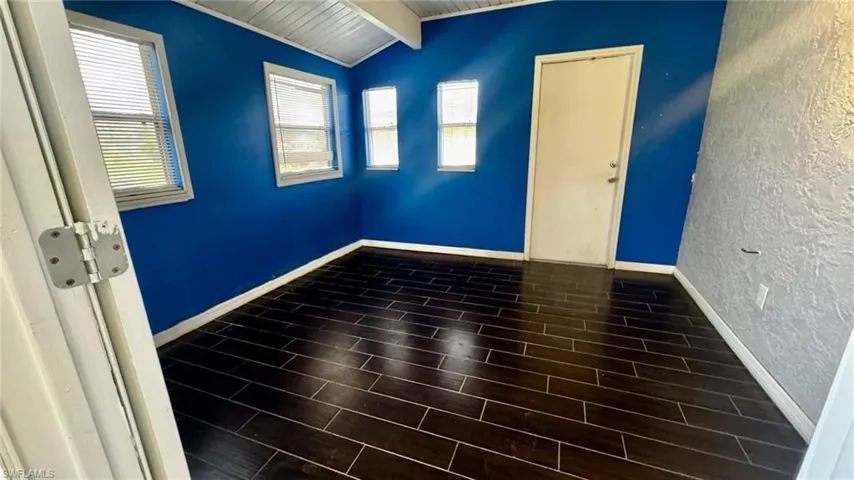 Interior room featuring dark wood-finish flooring, vaulted ceiling with exposed beams, textured accent wall, and multiple windows