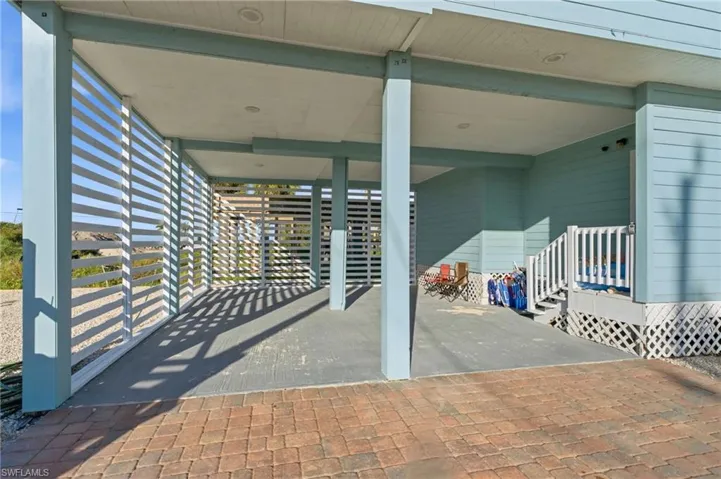 Covered ground-level parking area featuring concrete flooring, horizontal slat privacy screening, recessed lighting, and brick paver driveway - Virtually Edited Image
