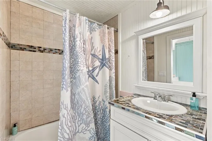Bathroom featuring a vanity with a stone-tile countertop, an integrated oval sink, beadboard wall paneling, tile shower surround with decorative glass accent inlay, and an overhead light fixture - Virtually Edited Image