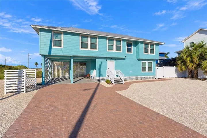 Elevated coastal home featuring horizontal lap siding, covered ground-level parking, a paved brick driveway, gravel surfacing, and white lattice skirting - Virtually Edited Image