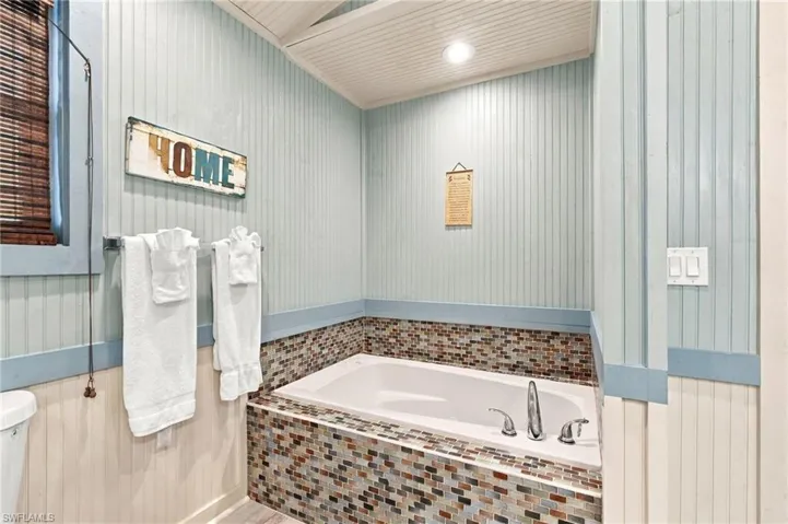 Drop-in soaking tub with a tiled surround featuring multicolor mosaic glass, vertical beadboard wall paneling, and recessed ceiling lighting - Virtually Edited Image