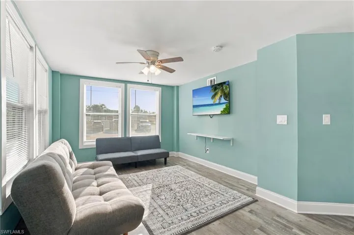 Living space featuring wood-finish flooring, dual windows, ceiling fan, and a wall-mounted floating shelf - Virtually Edited Image