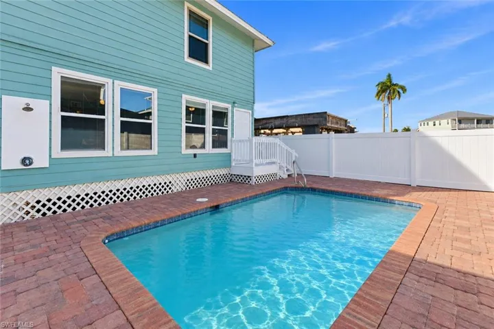 In-ground swimming pool featuring a brick paver deck, white privacy fencing, exterior wall-mounted shower fixture, and lattice skirting - Virtually Edited Image