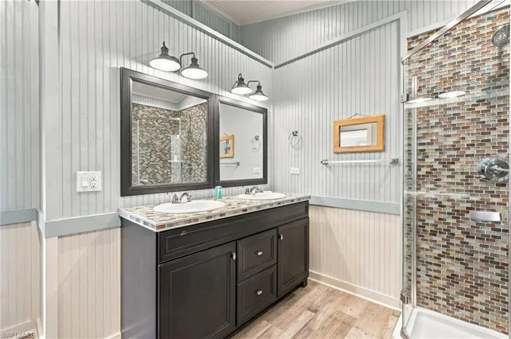 Bathroom featuring a dark wood-finish vanity with dual oval sinks, beadboard wall paneling, two-light fixtures, and a walk-in shower with multicolor mosaic tile surround - Virtually Edited Image
