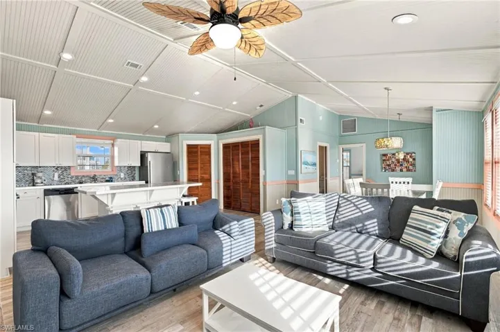 Open-concept living space featuring vaulted beadboard ceilings, recessed lighting, wood-finish flooring, and a kitchen with white cabinetry and a built-in peninsula - Virtually Edited Image