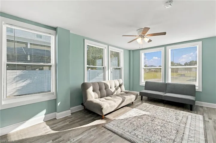 Interior room featuring wood-finish flooring, light-toned wall paint, multiple windows with grid detailing, and a ceiling-mounted fan with integrated lighting - Virtually Edited Image