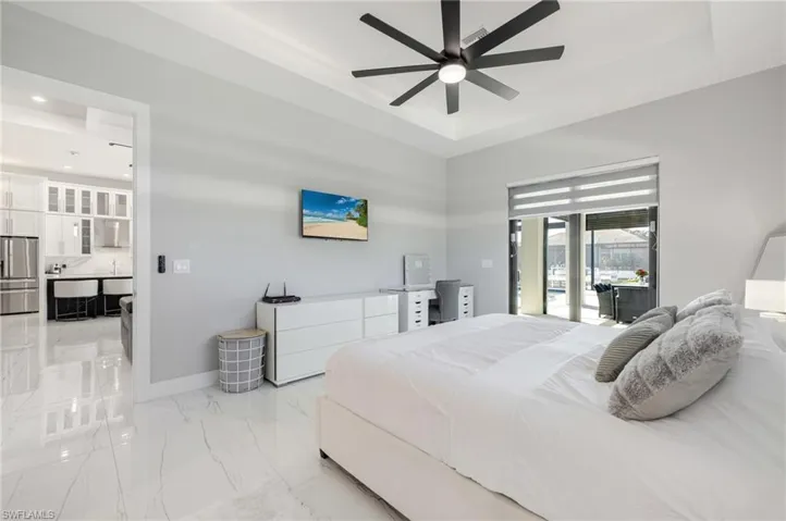 Bedroom featuring a tray ceiling, stainless steel refrigerator with ice dispenser, access to outside, light marble finish flooring, and a ceiling fan