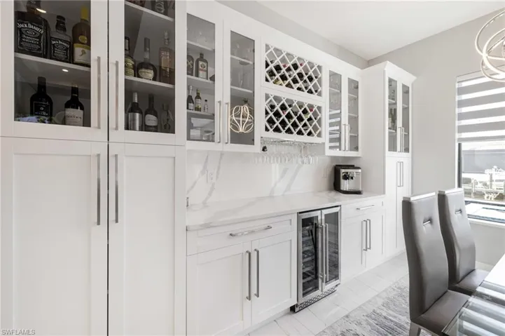 Indoor dry bar with beverage cooler, light stone countertops, white cabinets, glass fronted cabinets, and tasteful backsplash