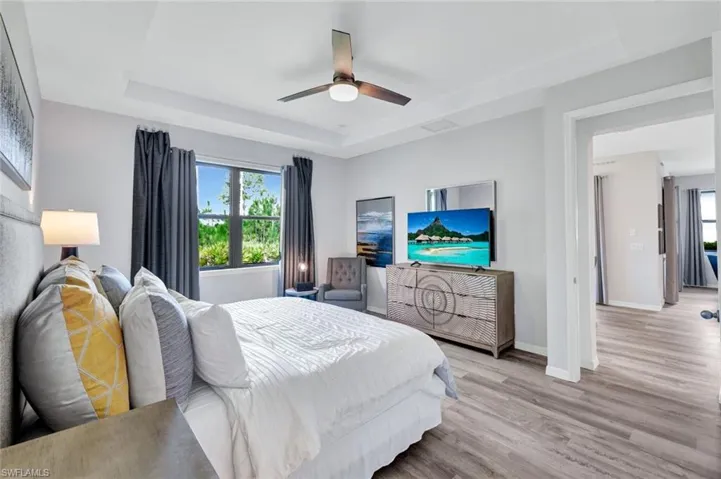 Spacious bedroom featuring wood-finish flooring, a tray ceiling with a modern ceiling fan, large window with outdoor scenery, and an open doorway leading to an additional space