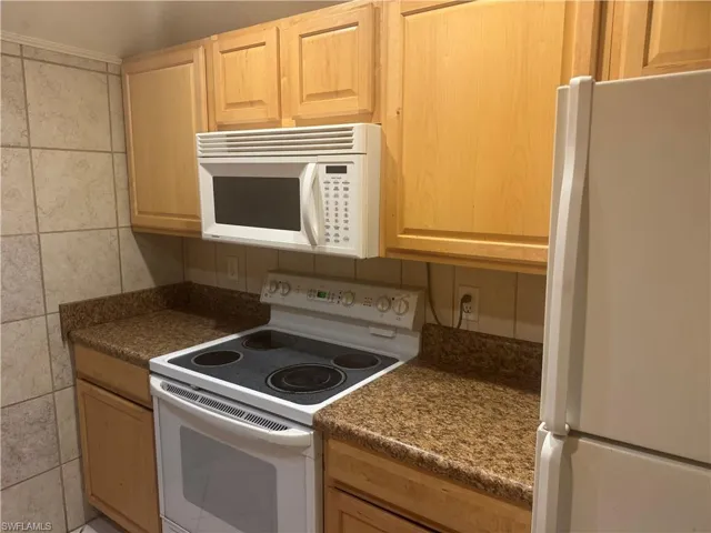 Kitchen with white appliances, dark countertops, light wood finish cabinets, and tile walls