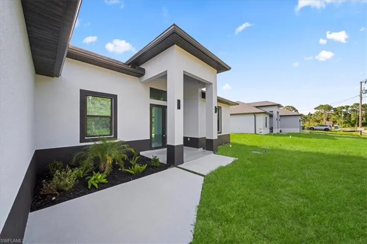 Modern exterior featuring a prominent covered entry, clean white stucco finish, dark trim accents, a large window, and mature landscaping