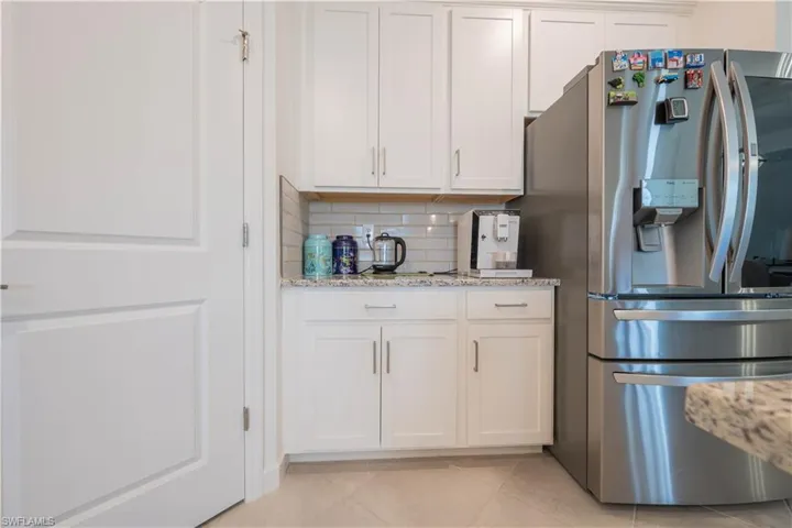 Kitchen with stainless steel fridge, backsplash, white cabinetry, light stone countertops, and light tile patterned flooring
