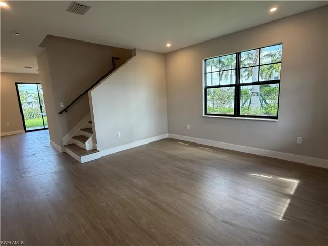 Spacious living area featuring light-toned flooring, recessed lighting, and a large window providing ample natural light and views of tropical foliage