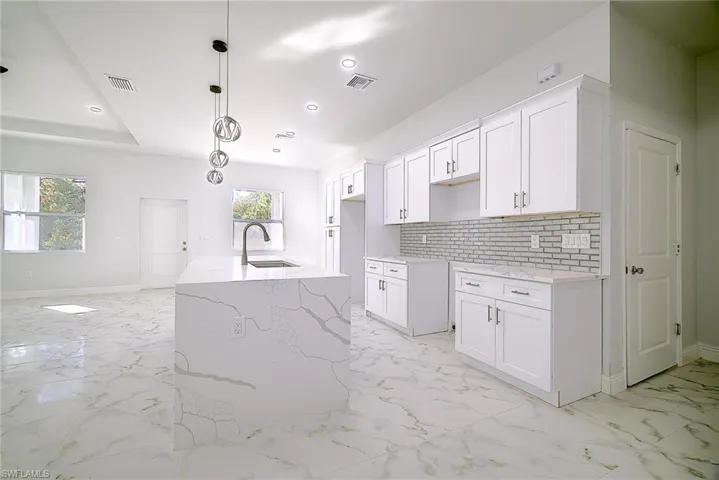 Kitchen featuring light stone counters, backsplash, white cabinetry, decorative light fixtures, and light marble finish flooring
