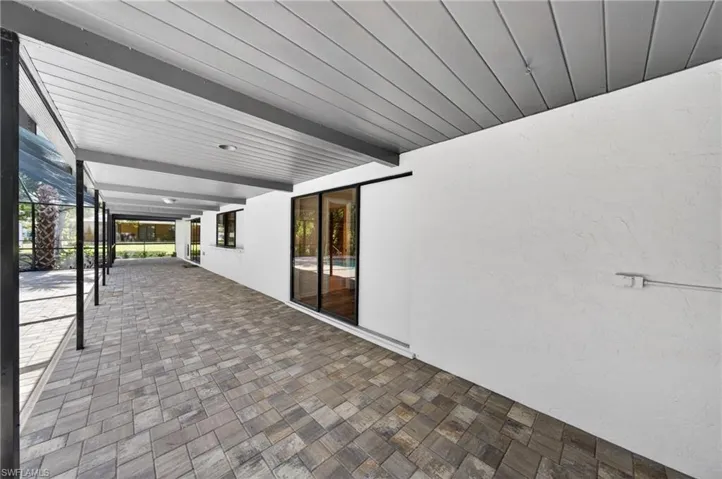 Covered outdoor walkway featuring textured pavers, an exposed beam ceiling, and multiple glass sliding doors providing access to the interior