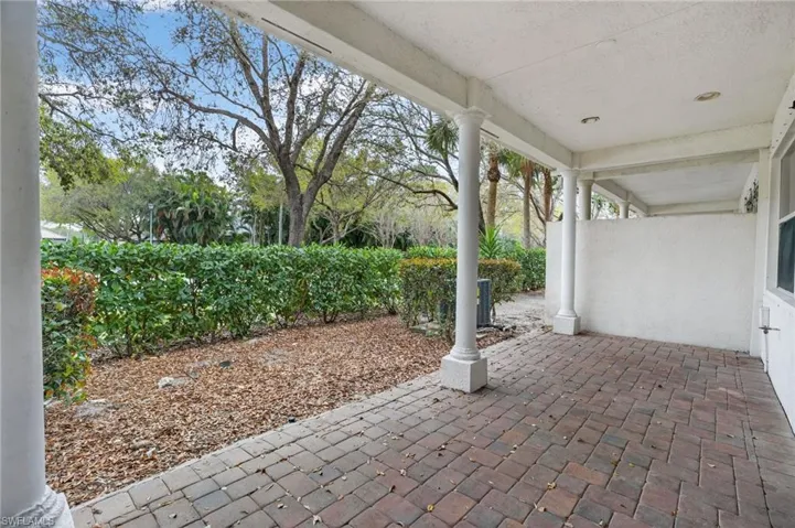 Covered outdoor patio featuring brick pavers, white columns, mature landscaping, and an exterior wall