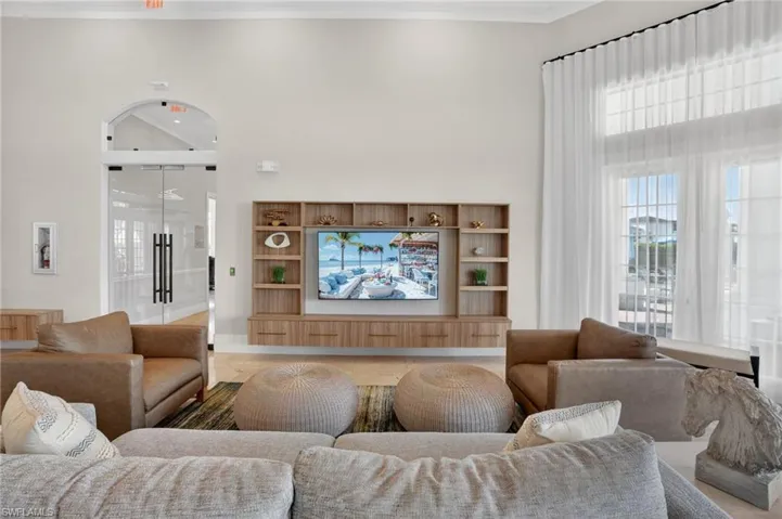 Spacious living area featuring a custom wood-finish media console with integrated shelving, an arched entryway with double glass doors, and tall windows with sheer drapery