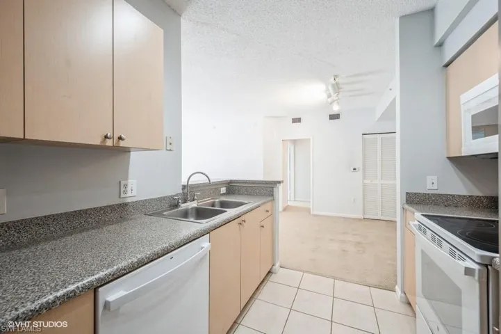 Kitchen with sink, white appliances, light carpet, light brown cabinets, and a textured ceiling