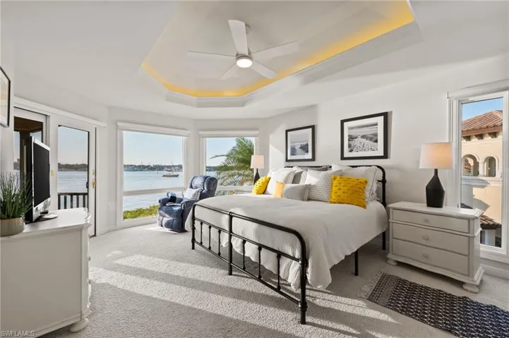 Carpeted bedroom with a raised ceiling, multiple windows, ceiling fan, and a water view