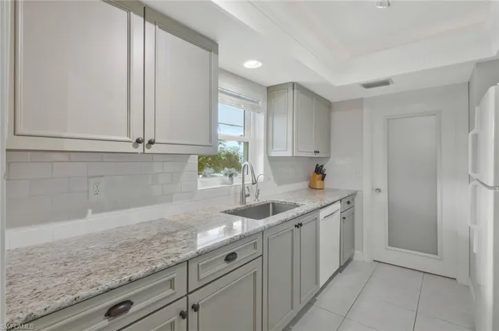 Kitchen featuring a tray ceiling, wood cabinetry, granite countertops, subway tile backsplash, tile flooring and white appliances.  Laundry room/storage through the door by the refrigerator.