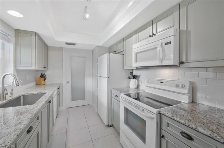 Kitchen featuring a tray ceiling, wood cabinetry, granite countertops, subway tile backsplash, tile flooring and white appliances.  Laundry room/storage through the door by the refrigerator.