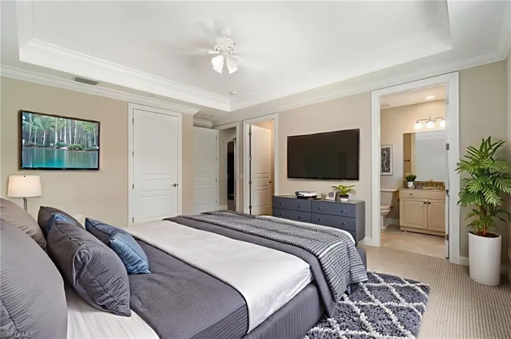 Bedroom with a raised ceiling, light colored carpet, crown molding, ensuite bath, and baseboards