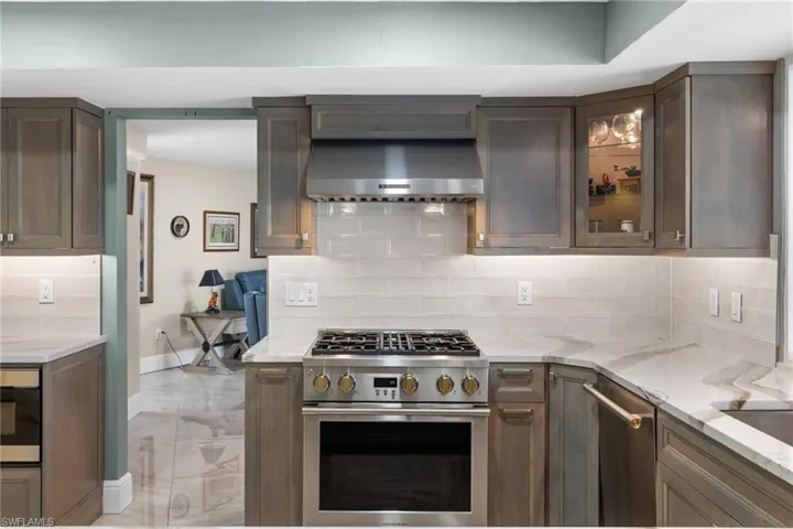 Kitchen with light stone counters, stainless steel appliances, decorative backsplash, glass insert cabinets, and wall chimney range hood