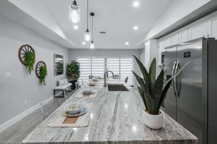Kitchen with freestanding refrigerator, an island with sink, light stone counters, decorative light fixtures, and white cabinetry