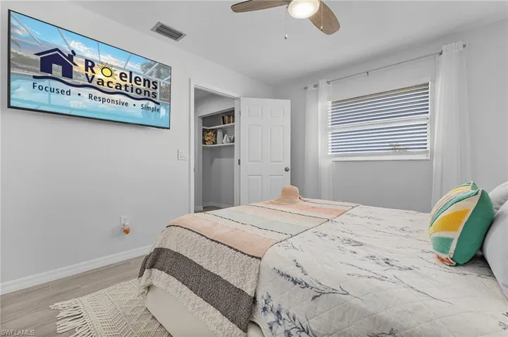 Recessed wall-mounted television, ceiling fan with integrated lighting, wood-finish flooring, and a window with blinds and sheer curtains