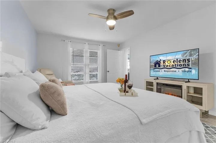Bedroom featuring a ceiling fan with integrated lighting, window with horizontal blinds and sheer curtains, a light-toned wood-finish media console, and a built-in electric fireplace