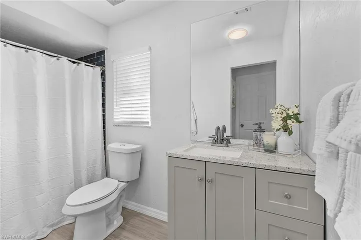 Bathroom featuring a gray vanity with a light-toned countertop, a large wall-mounted mirror, wood-finish flooring, a toilet, and a shower with dark subway-style tile