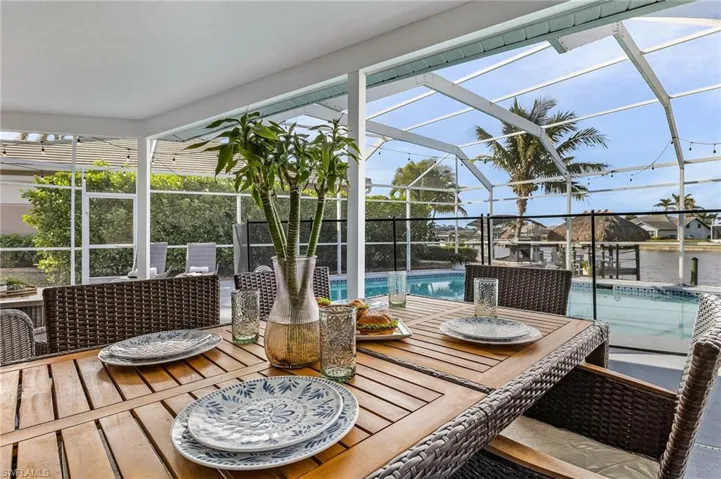 Screened lanai featuring a wood-finish dining table and woven chairs, an in-ground pool, and a canal with a thatched-roof boat house
