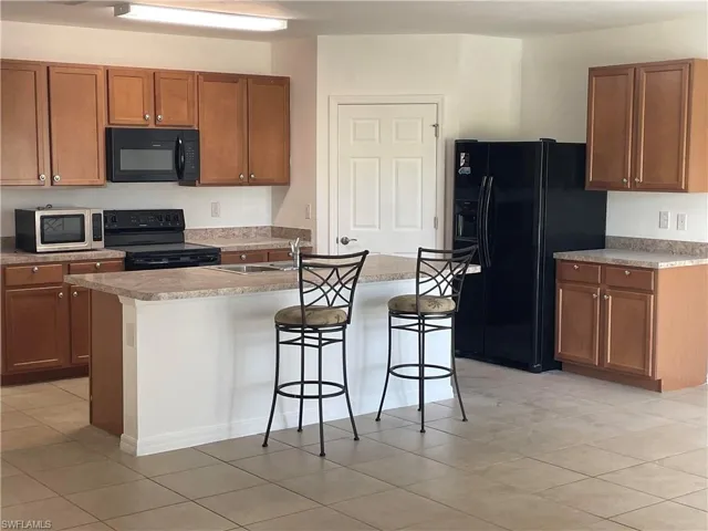 Kitchen with brown cabinetry, black appliances, a breakfast bar, an island with sink, and light tile patterned floors