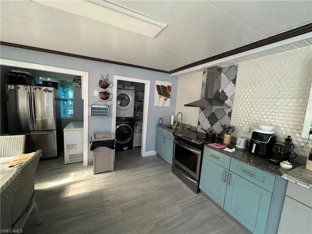 Kitchen featuring appliances with stainless steel finishes, wall chimney exhaust hood, stacked washer and clothes dryer, tasteful backsplash, and dark stone counters