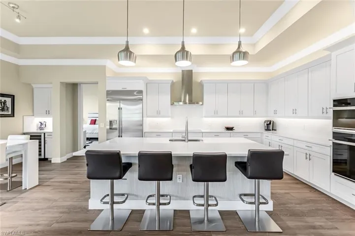 Kitchen featuring a center island with sink, built in fridge, a tray ceiling, white cabinetry, and wall chimney exhaust hood