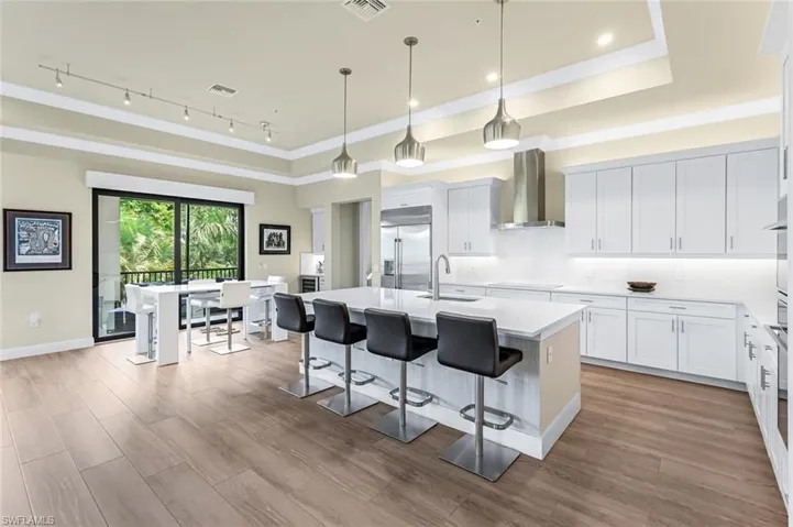 Kitchen featuring wall chimney range hood, light wood-type flooring, an island with sink, stainless steel built in refrigerator, and a raised ceiling